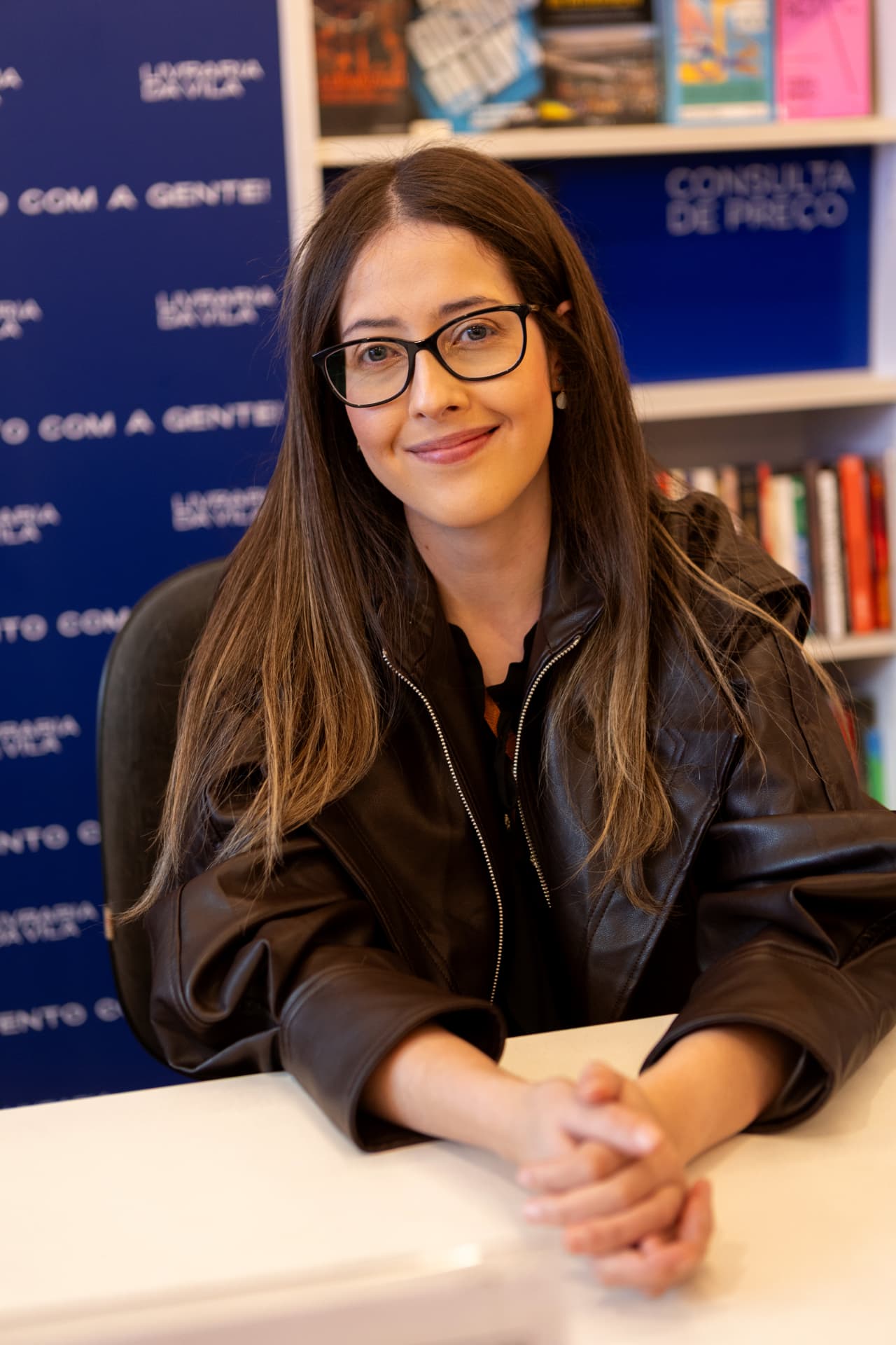 Joice Cristina sentada em uma mesa de autógrafos, sorrindo